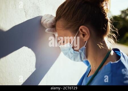 coronavirus pandemic. stressed modern medical practitioner woman in uniform with medical mask and rubber gloves outdoors in the city near wall. Stock Photo