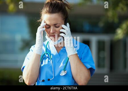 coronavirus pandemic. stressed modern physician woman in scrubs with stethoscope and medical mask talking on a smartphone outside near clinic. Stock Photo
