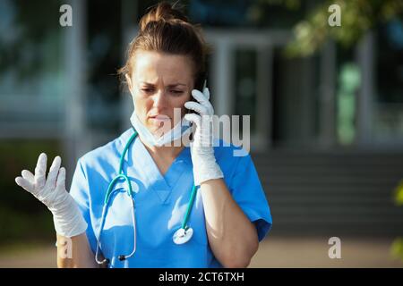 coronavirus pandemic. unhappy modern physician woman in uniform with stethoscope and medical mask using a smartphone outdoors near clinic. Stock Photo