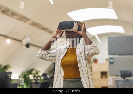Waist up portrait of young African-America woman wearing VR gear in office and enjoying immersive reality, copy space Stock Photo