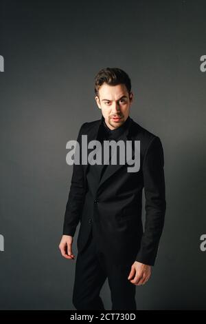 Young handsome man in a black suit, standing and seriously looking at the camera, against plain studio background Stock Photo