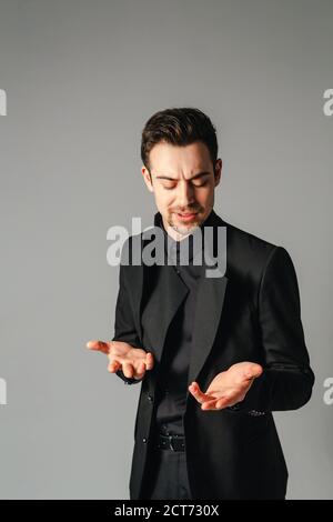 Close up portrait of a young handsome man in a black suit, standing, seriously looking at his hands while explaining something, against plain studio b Stock Photo