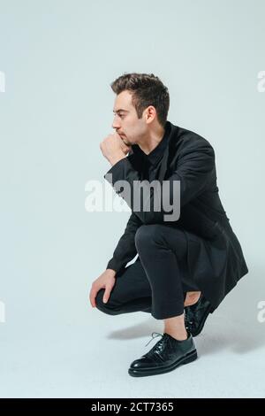 Portrait of a young handsome man in a black suit, seriously looking to the side, hand near face, against plain studio background Stock Photo