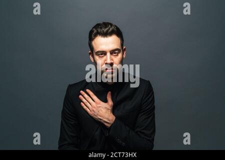 Close up portrait of a young handsome man in a suit, hand flat on the chest, looking at the camera, against plain studio background Stock Photo
