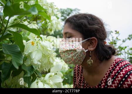 Beautiful lady with floral mask, earrings and designer top smells white Mussaenda flowers. Protection from corona in a stylish manner. Stock Photo