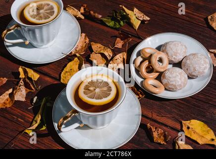 Two white ceramic cups with beautiful gold shiny handles with tea and lemon. Snacks and sweets for tea on a small white saucer. Autumn leaves and tea Stock Photo