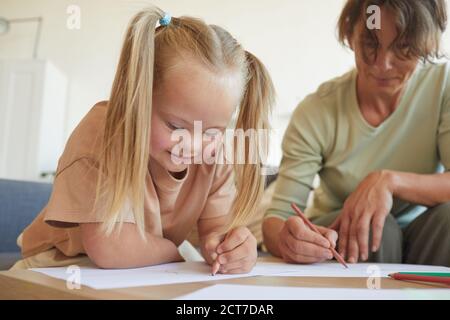 Portrait of cute blonde girl with down syndrome drawing with mother or teacher while enjoying development exercises Stock Photo
