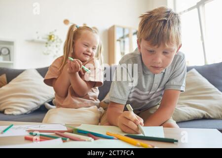 Portrait of cute blonde girl with down syndrome laughing happily while watching brother drawing at home, copy space Stock Photo