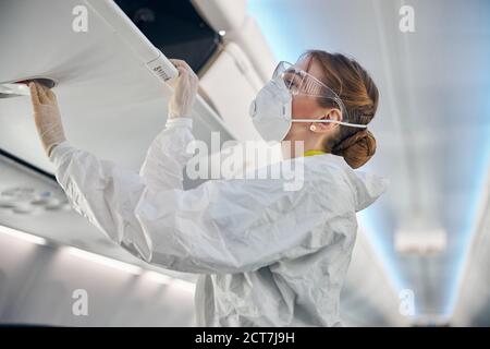Beautiful woman flight attendant in mask and glasses closing luggage compartment Stock Photo