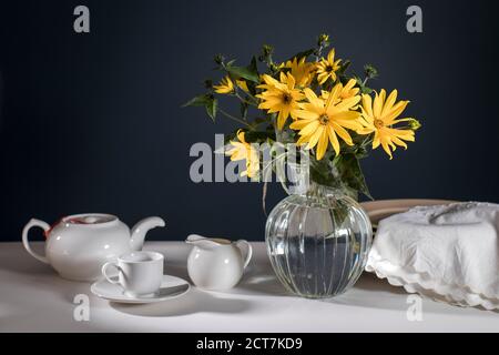 A bouquet of yellow jerusalem artichoke in a transparent vase on against black background on the table. White teapot, cup and saucer and milk jug for Stock Photo