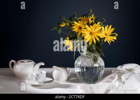 A bouquet of yellow jerusalem artichoke in a transparent vase on against black background on the table. White teapot, cup and saucer and milk jug for Stock Photo