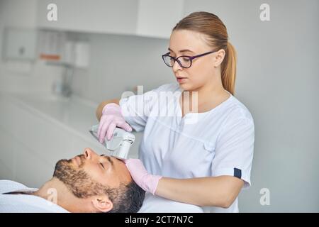 Charming female esthetician treating male skin with laser device Stock Photo