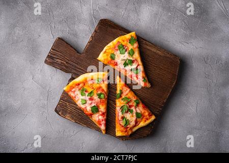 Hot pizza slices with mozzarella cheese, ham, tomato and parsley on brown wooden cutting board, stone concrete background, top view Stock Photo