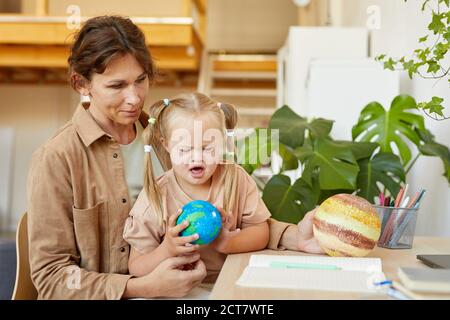 Portrait of cute girl with down syndrome holding planet model while studying at home with mother , copy space Stock Photo