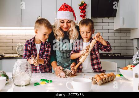 Happy family of mother and two children twins sons bake cookies in kitchen before Christmas in decorated light kitchen Stock Photo