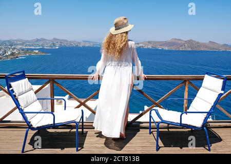 Beautiful woman from back in swimsuit white dress and straw hat on white terrace balcony of house or hotel with Sea View Stock Photo