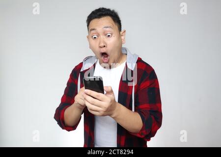 Attractive young Asian man reading texting chatting  on his phone, shocked surprised expression Stock Photo