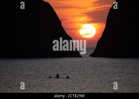 Three silhouetted unidentifiable surfers return from the sea in front of Gull Rocks at Holywell Bay in Cornwall as the sun sets behind the clouds. Stock Photo