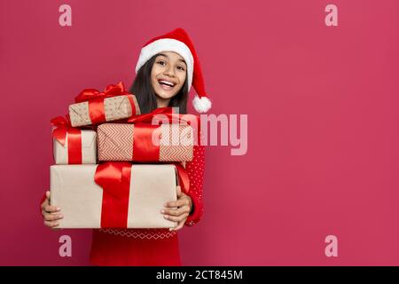 Excited indian kid girl wear santa hat hold many gifts boxes on red background. Stock Photo