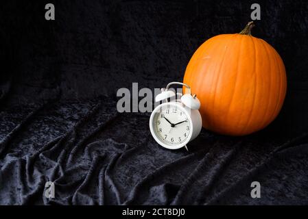 Orange pumpkin and classic white alarm clock on a black velvet background, fall time change Stock Photo