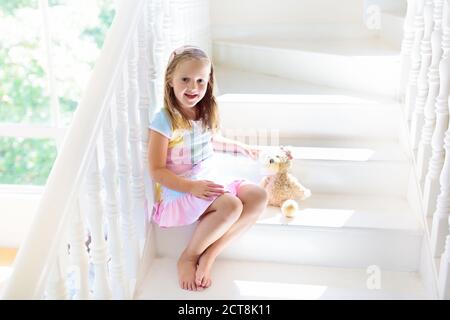 Kid walking stairs in white house. Little girl playing in sunny staircase. Family moving into new home. Child climbing steps of modern stairway. Foyer Stock Photo