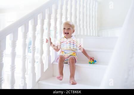 Kid walking stairs in white house. Baby boy playing in sunny staircase. Family moving into new home. Child crawling steps of modern stairway. Foyer an Stock Photo