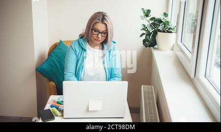 Front view photo of a busy caucasian senior lady with blonde hair working at the laptop from home near the window Stock Photo