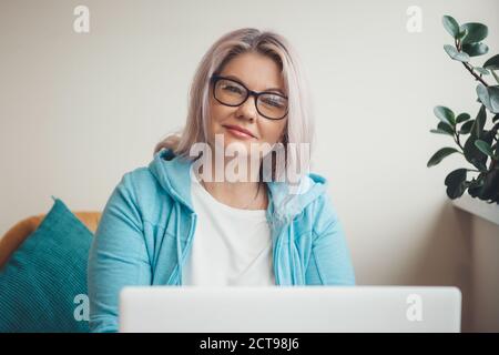 Front view photo of a senior caucasian woman looking at camera through eyeglasses and smile while using a laptop and working remotely Stock Photo