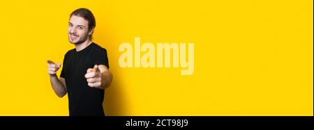 Bearded caucasian man in a black t-shirt pointing at camera and smile on a yellow studio banner Stock Photo