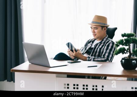 Happy Asian young man sitting at his table reading at home office, Stay home. quarantine. Stock Photo