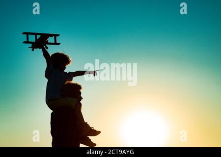 Sunset silhouette of Father carrying his son on shoulders. Child son pilot and dad daddy father with airplane. Stock Photo