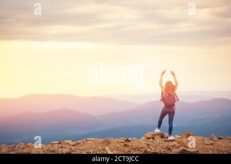 Happy girl stands on top of mountain, raises hands up against background of sunset, photograph from back Stock Photo