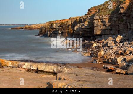 A long exposure picture of incoming waves crashing onto the rock face and boulders on a small bay beach on a sunny day. Stock Photo