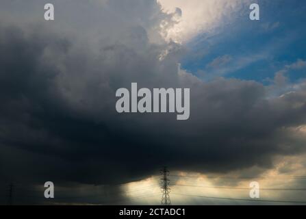 Blue sky and gray clouds with white sunlight above high voltage electric pylons and transmission lines. Sun rays through dark clouds. High voltage Stock Photo