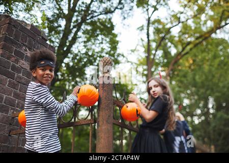 Portrait of kids dressed in Halloween costumes holding onto fence while playing with friends outdoors, copy space Stock Photo