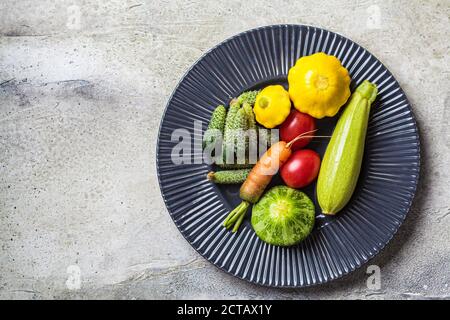 Mini vegetables concept. Mini zucchini, tomatoes, cucumbers, squash, and carrots on a dark plate. Stock Photo
