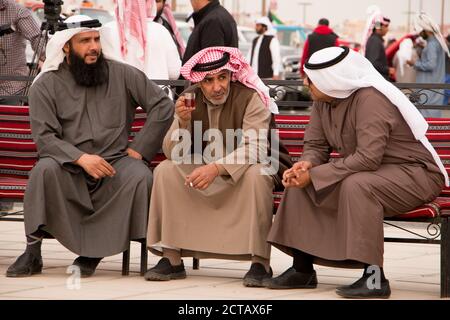 Three Kuwaiti men talking and smoking on a bench. Dressed in traditional winter colored dishdashas. Kuwait, Middle East. Stock Photo