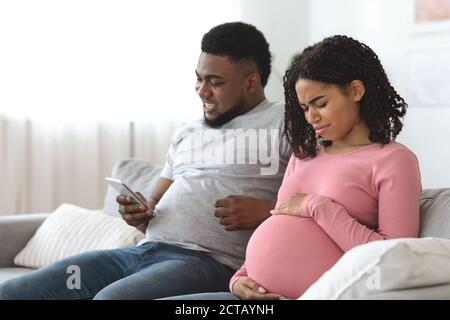 Pregnant black woman suffering from pain while husband using phone Stock Photo