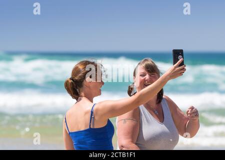 Women on a staycation holiday laughing as they take a selfie with a smart phone at Fistral Beach in Newquay in Cornwall. Stock Photo