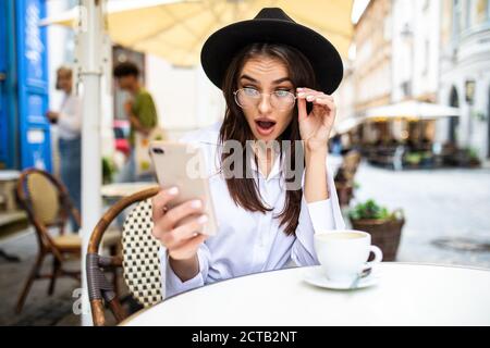 Shocked news. Cafe with wifi. Young amazed woman using smartphone while sitting sidewalk restaurant. Stock Photo