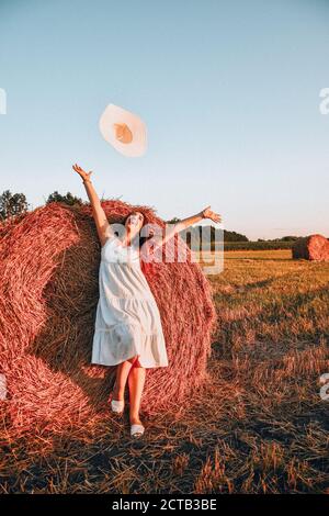 Portrait of young happy woman flying hat on the haystack in morning sunlight, countryside. Beautiful woman in a dress sits on a haystack. Cottagecore Stock Photo