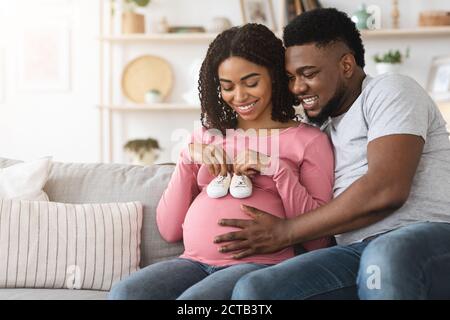 Happy black pregnant couple placing baby shoes on woman belly Stock Photo