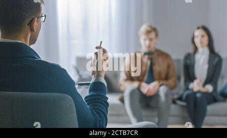 Unfocused Couple on Counseling Session with Psychotherapist. Focus on Back of Therapist Taking Notes and Talking: People Sitting on Analyst Couch Stock Photo