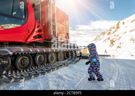 Cute adorable playful happy toddler boy play at red modern snowcat ratrack snowplow box grooming standing on peak alpine skiing resort Ischgl Austria Stock Photo