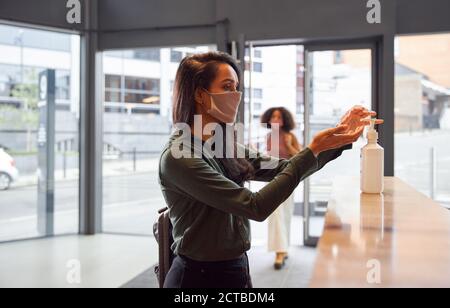 Mixed Race Businesswoman In Face Mask Uses Hand Sanitiser In Office Reception During Health Pandemic Stock Photo