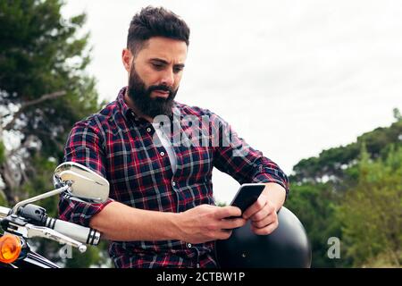 handsome bearded man consulting his mobile phone sitting on his motorcycle with his helmet under his arm, concept of freedom and biker lifestyle, copy Stock Photo