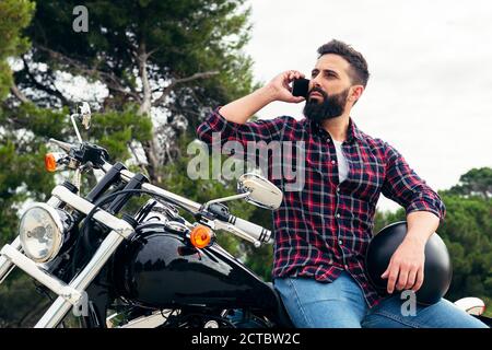 handsome bearded man talking on his mobile phone sitting on his motorcycle with his helmet under his arm, concept of freedom and biker lifestyle, copy Stock Photo