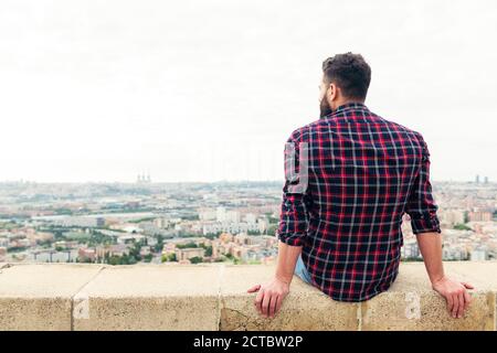 rear view of a bearded man sitting on the rooftop observing the city at his feet, concept of freedom and disconnection, copy space for text Stock Photo