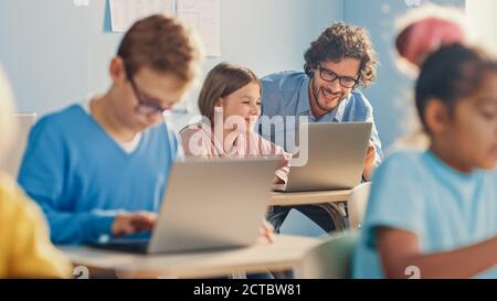 Elementary School Computer Science Class: Smart Girl Uses Laptop Computer, Friendly Teacher Helps Her by Explaining Lesson and Asignment. Children Stock Photo