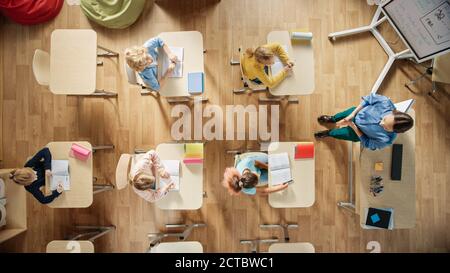 Bright Elementary School Classroom: Children Sitting at their School Desk Working, Doing Assignment, Enthusiastic Teacher Stands a the Head of the Stock Photo
